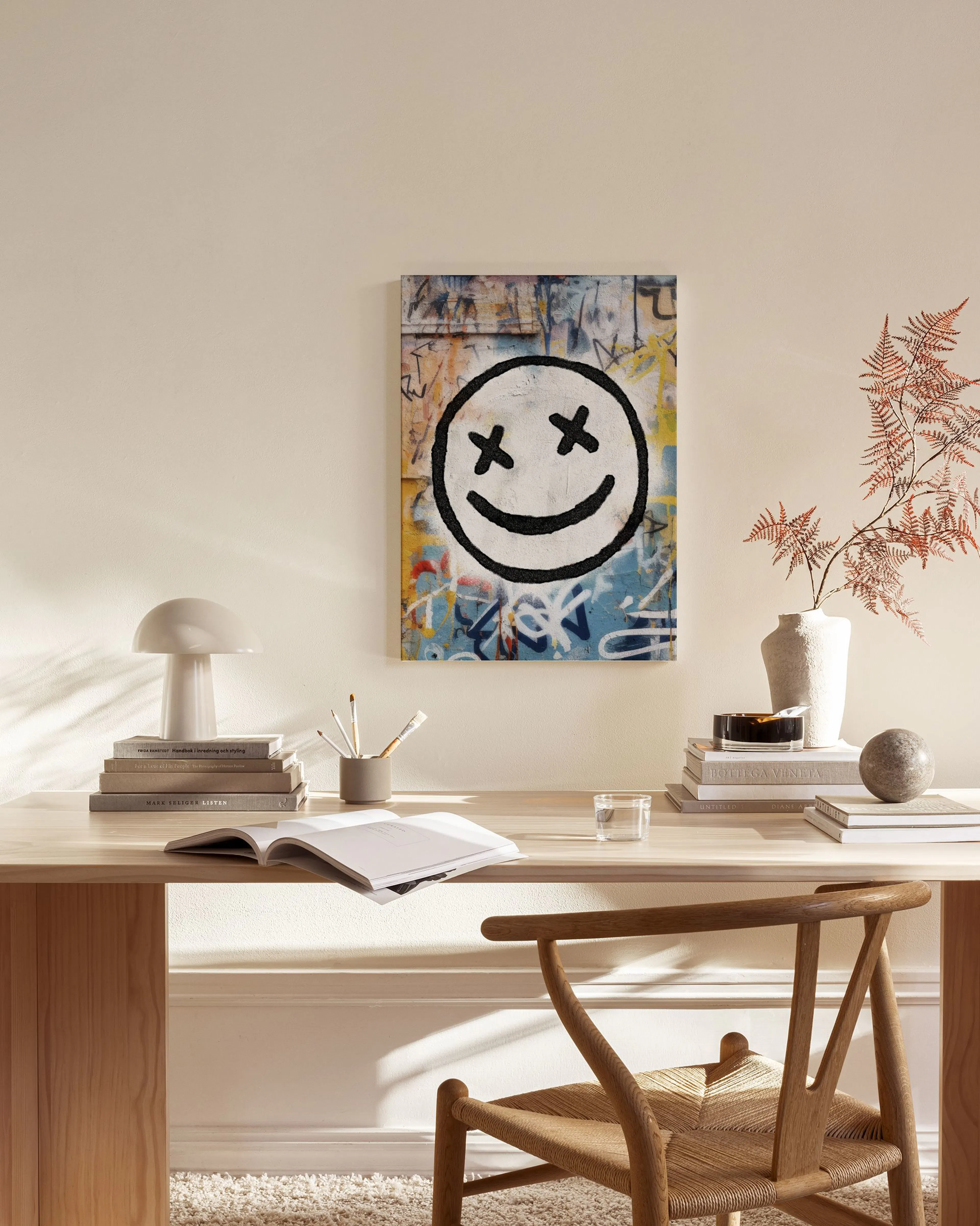 Lærred med graffiti-inspireret baggrund og en sort smiley med krydsede øjne, hængende i en stue over en træbænk.