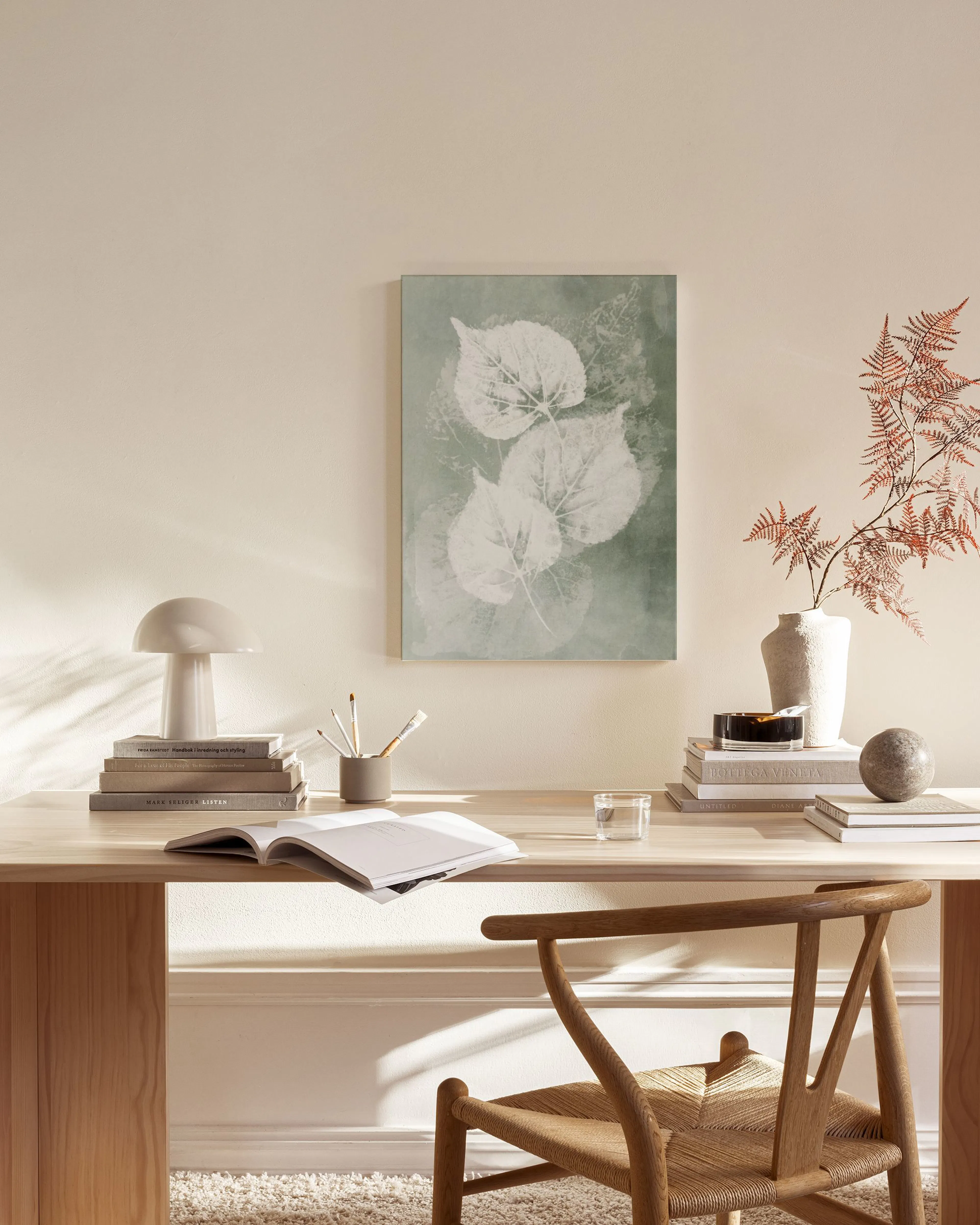 A botanical print featuring three white leaf skeletons on a sage green background, displayed above a wooden console table in a m