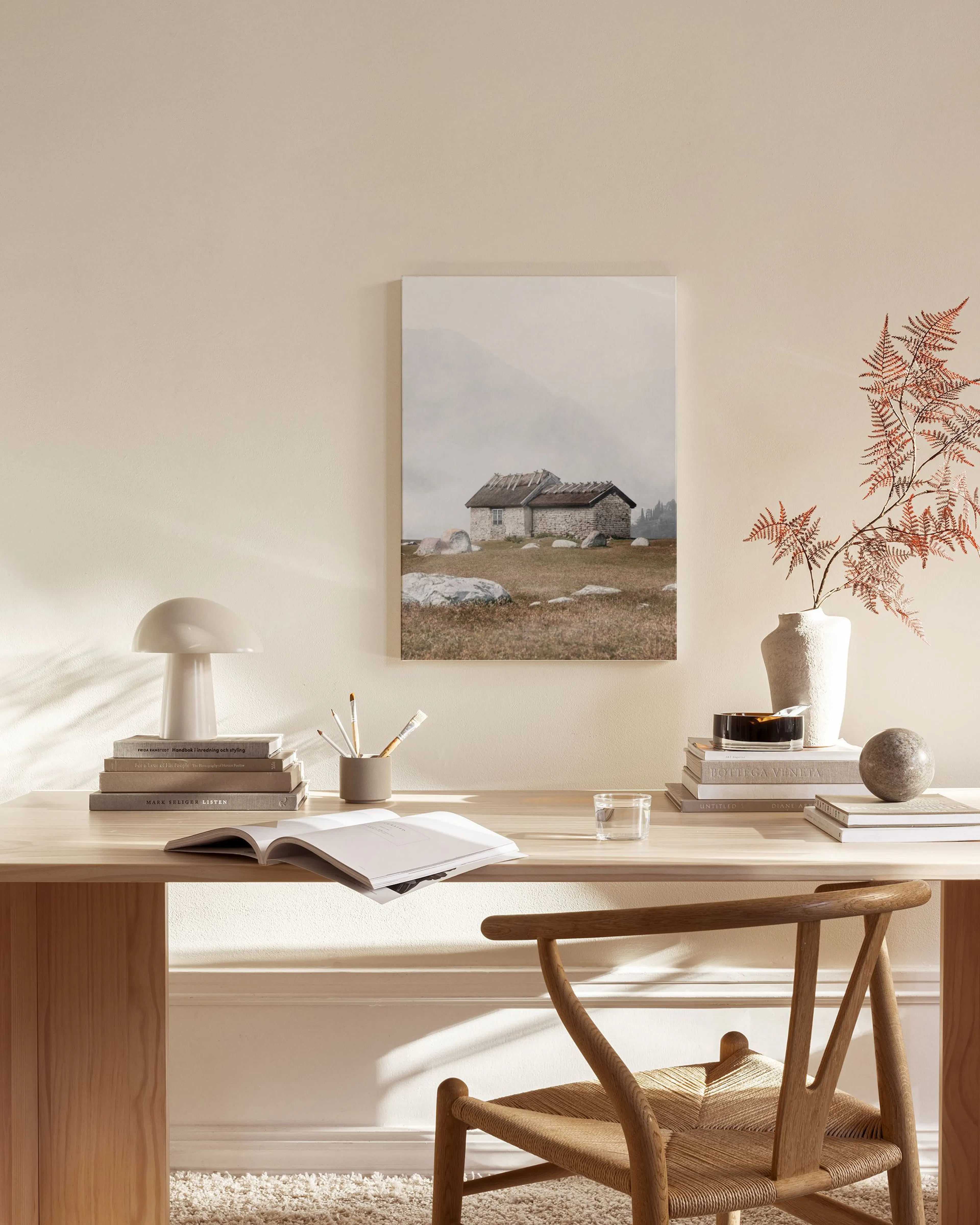 Toile de chalet en pierre dans un paysage montagneux brumeux, accrochée dans un salon moderne avec fauteuil en bois et table bas