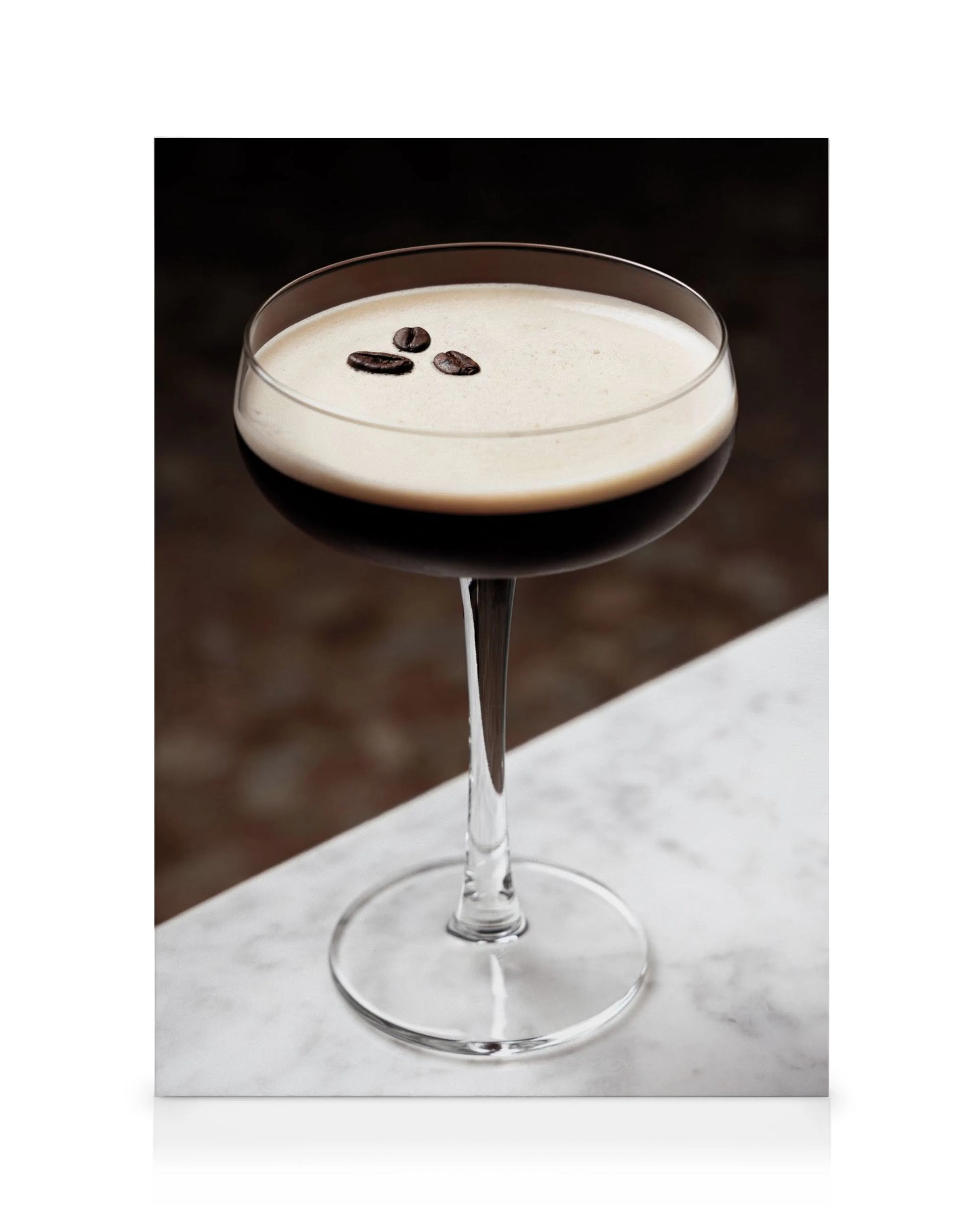 Bebida de café oscuro en vaso alto con espuma blanca y tres granos de café, sobre mármol blanco y fondo marrón.