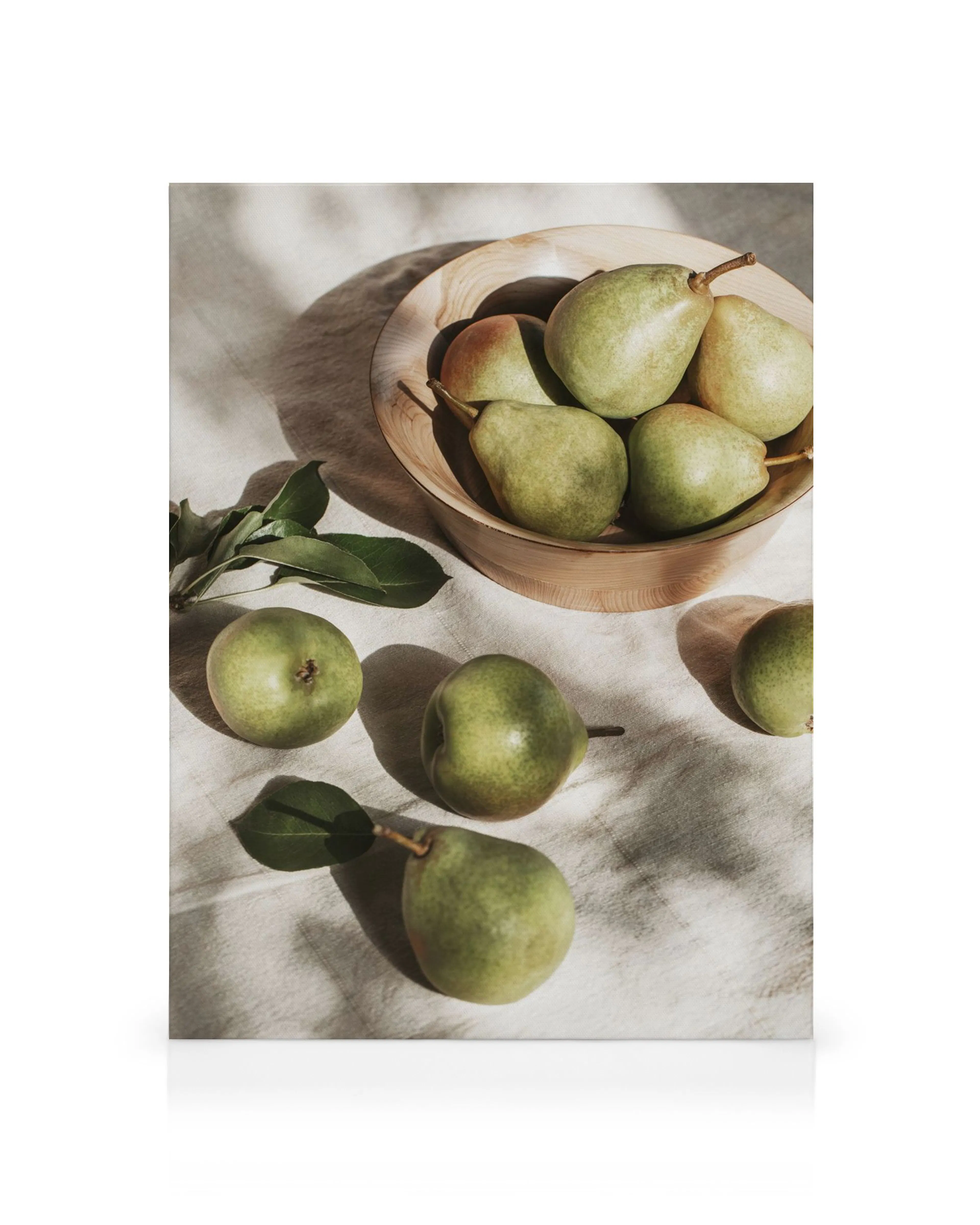 Sunlit pears in a wooden bowl with scattered fruit and green leaves on a beige cloth.