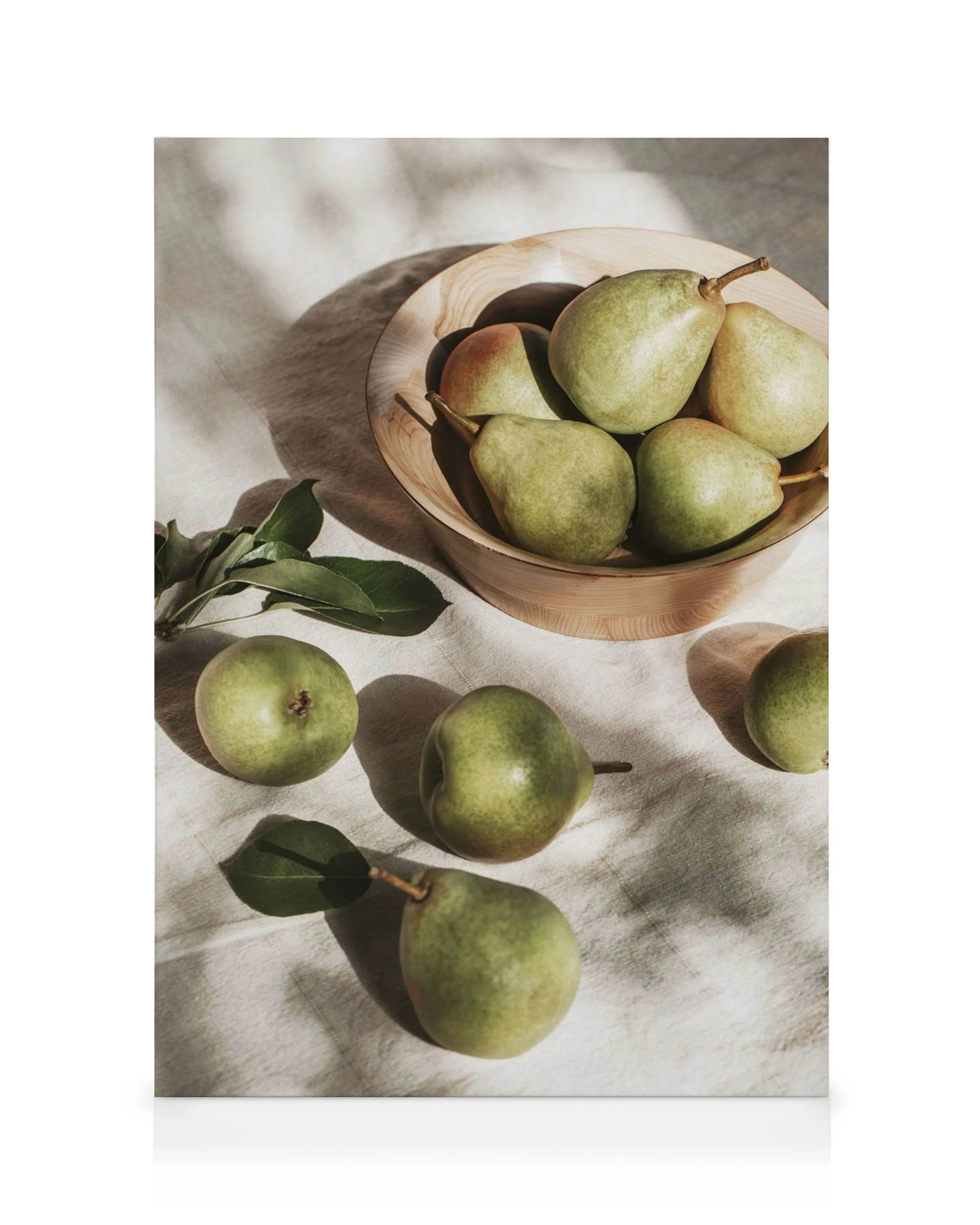 Sunlit green pears in a wooden bowl with scattered fruit and leaves on a beige cloth, creating a serene still life.