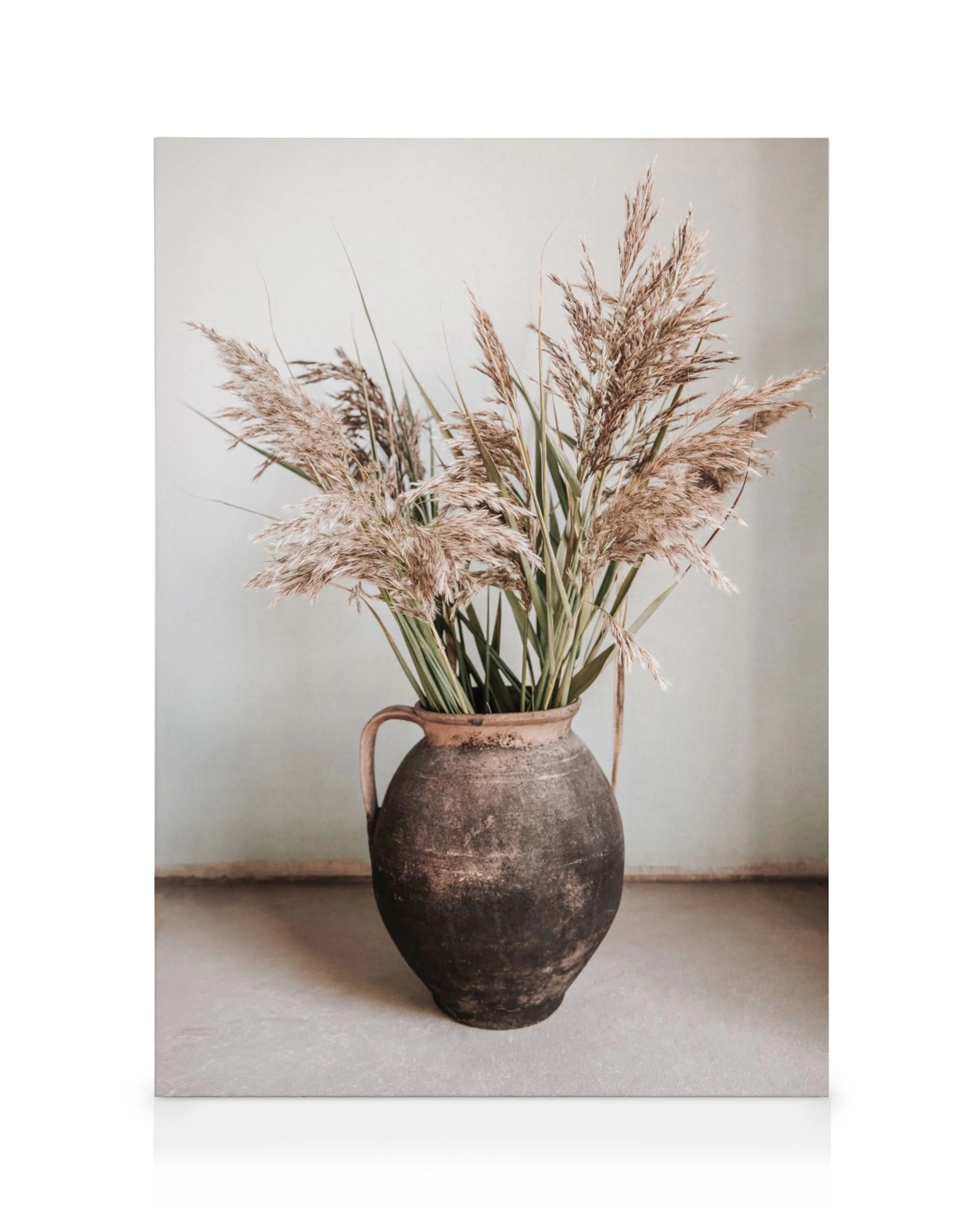 Leinwand mit rustikaler Tonvase, gefüllt mit hohen Schilfrohren und grünen Blättern auf grauem Boden vor blassgrüner Wand. Natur