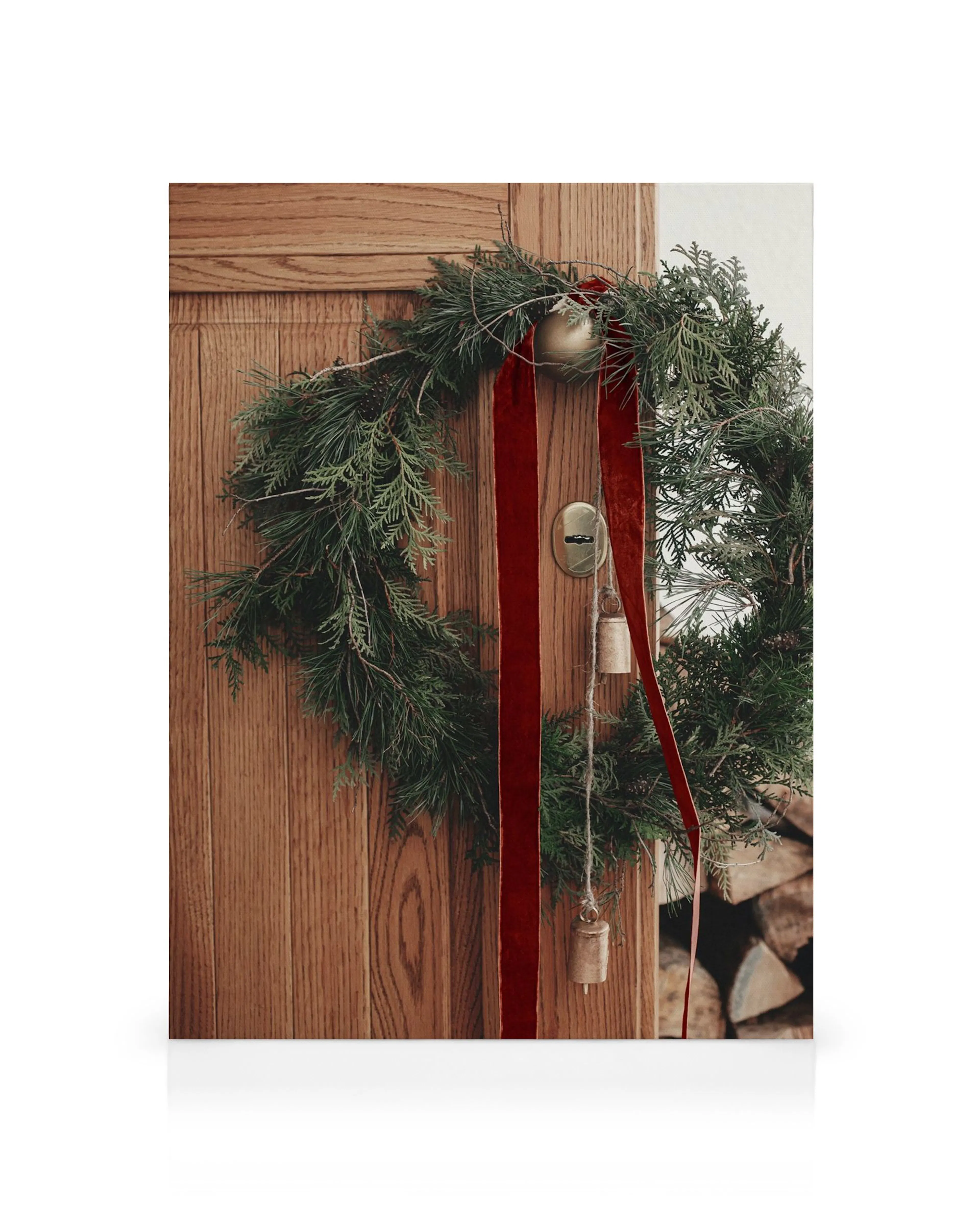 Rustic green holiday wreath with red ribbon on a wooden door, featuring bells and an ornament.