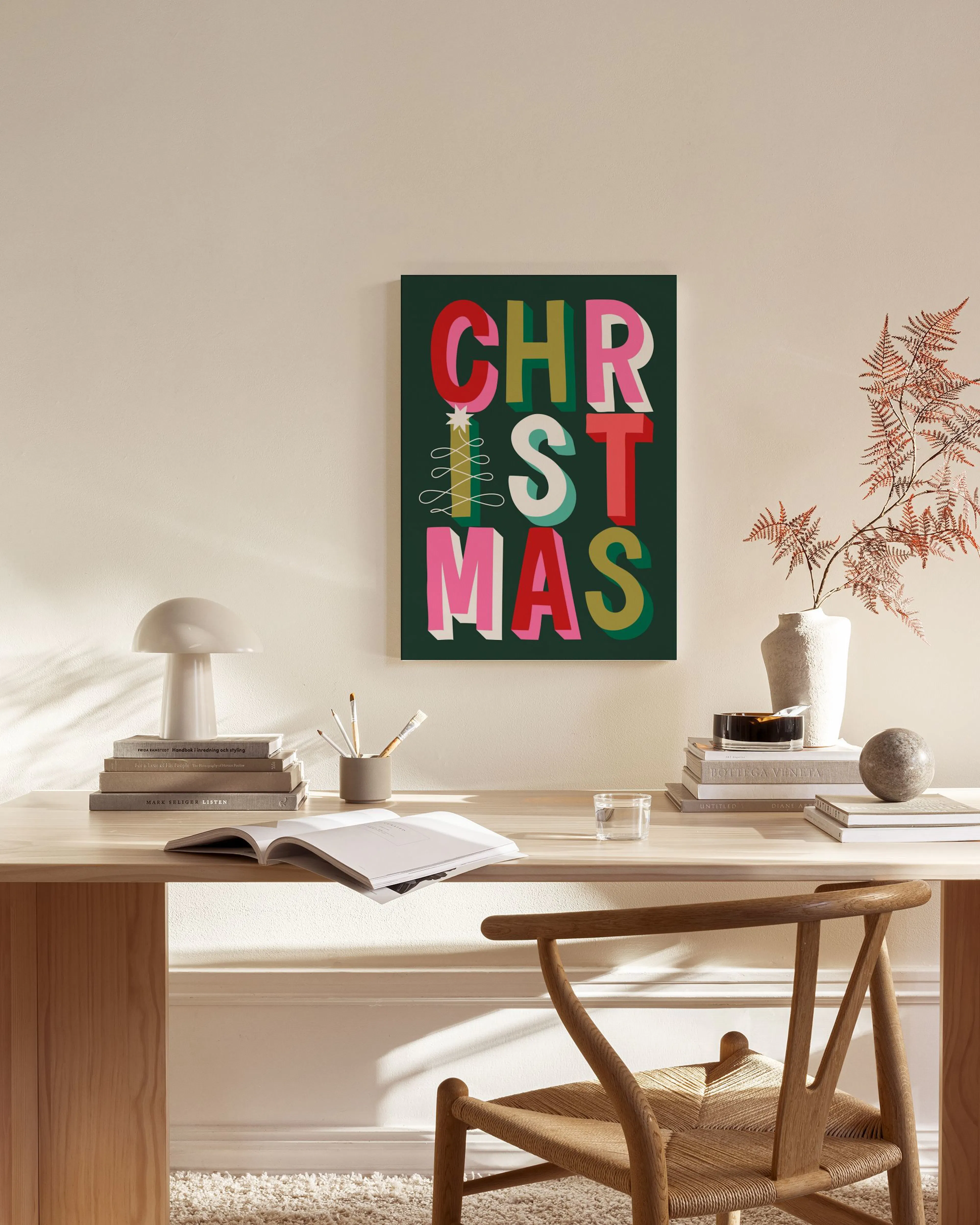 Lærred med ordet CHRISTMAS i grønne, røde, lyserøde og gule farver, hængende i en stue over et træmøbel.