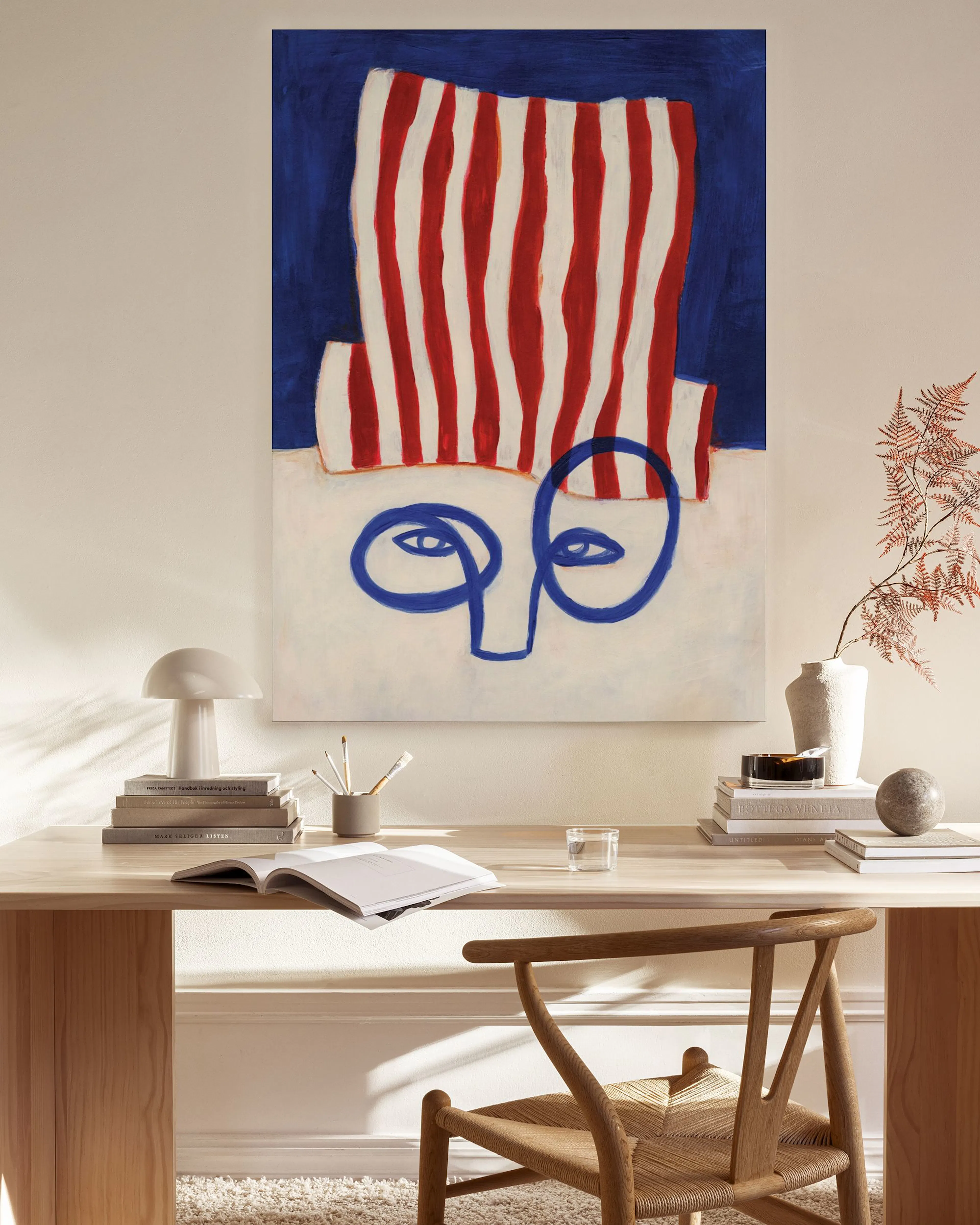 A canvas print of an abstract face with a red and white striped hat on a blue background, hanging in a minimalist living room.
