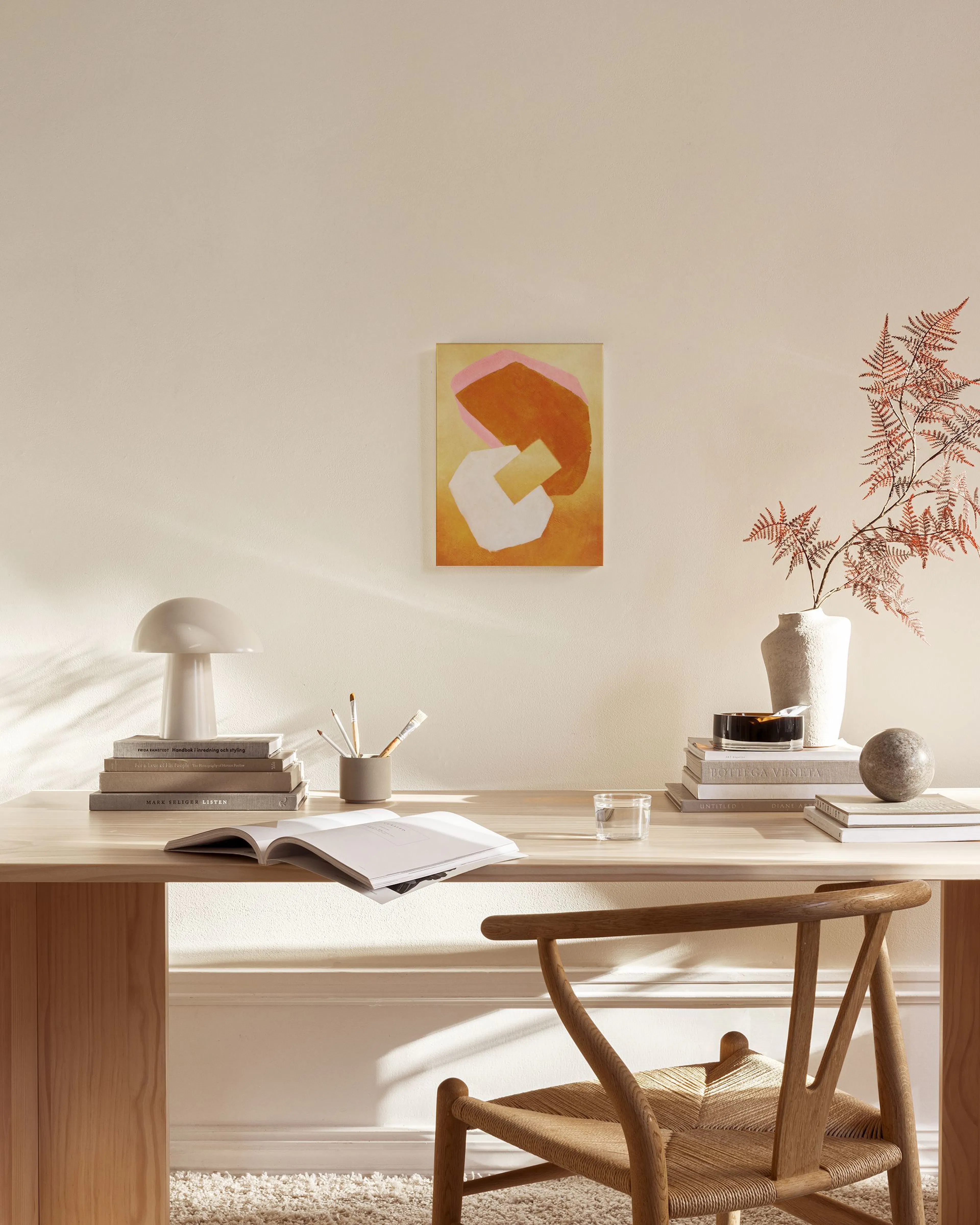 Tela decorativa com formas orgânicas laranjas, rosa e brancas sobre fundo amarelo, pendurada numa sala de estar minimalista.
