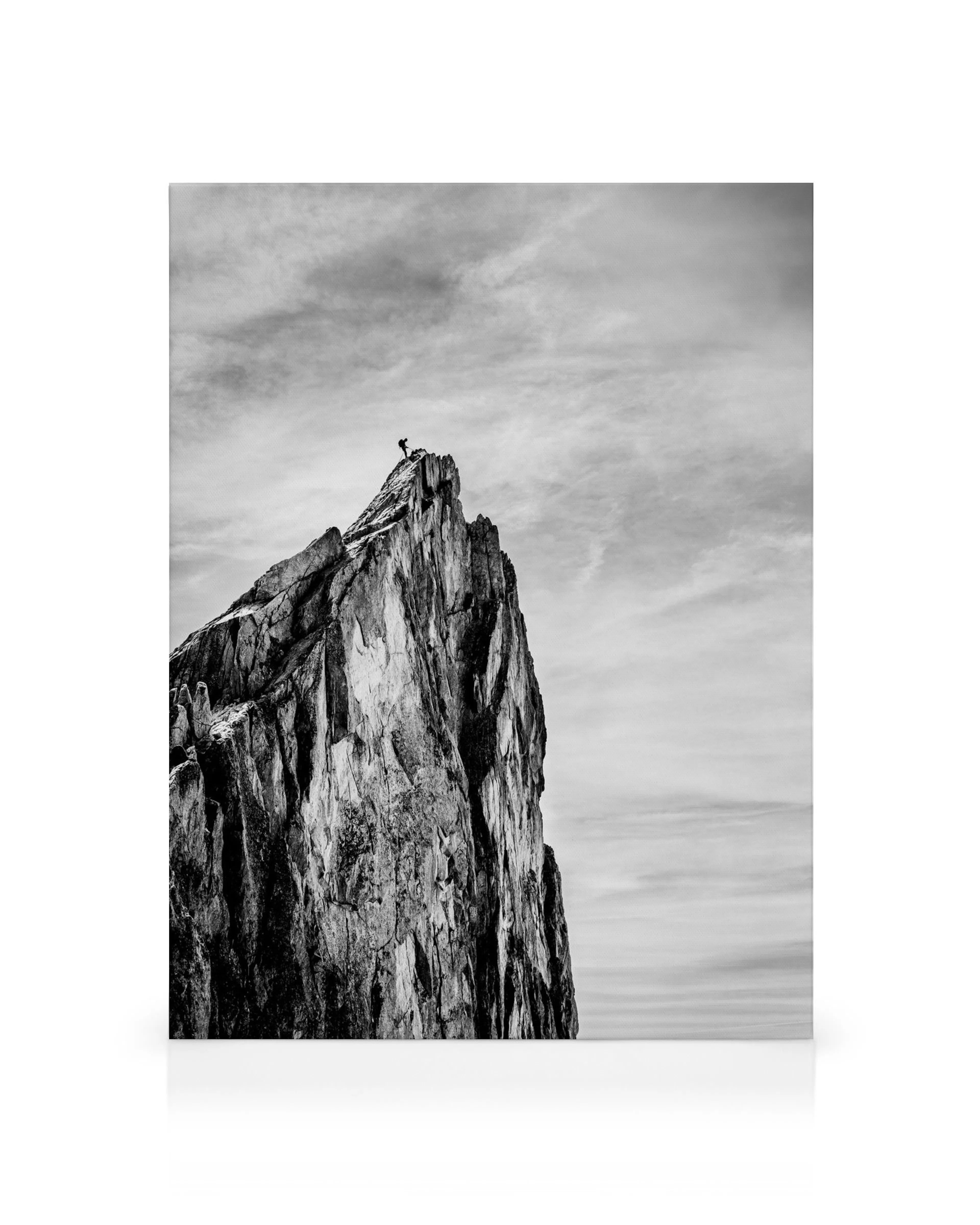 Black and white canvas print of a person standing on a jagged mountain peak under cloudy skies, black white photography.