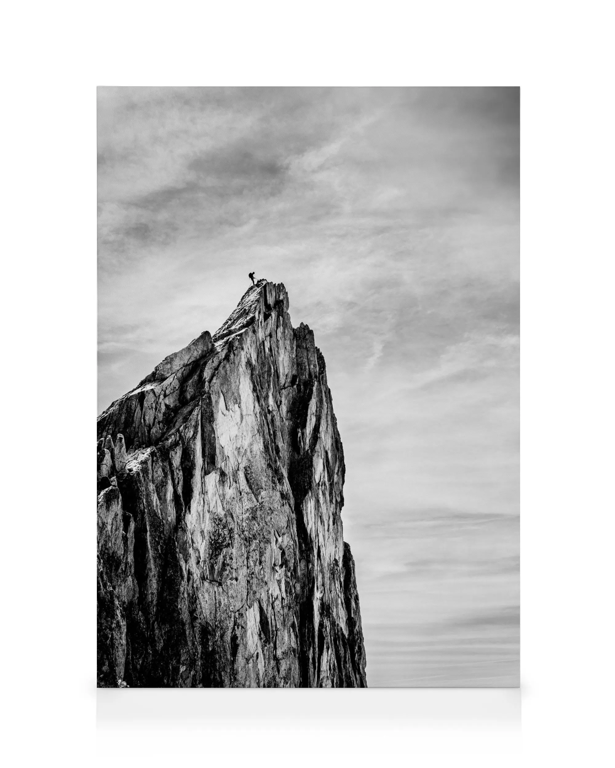 Black and white photography of a solitary figure standing atop a jagged mountain peak, reaching for the sky.