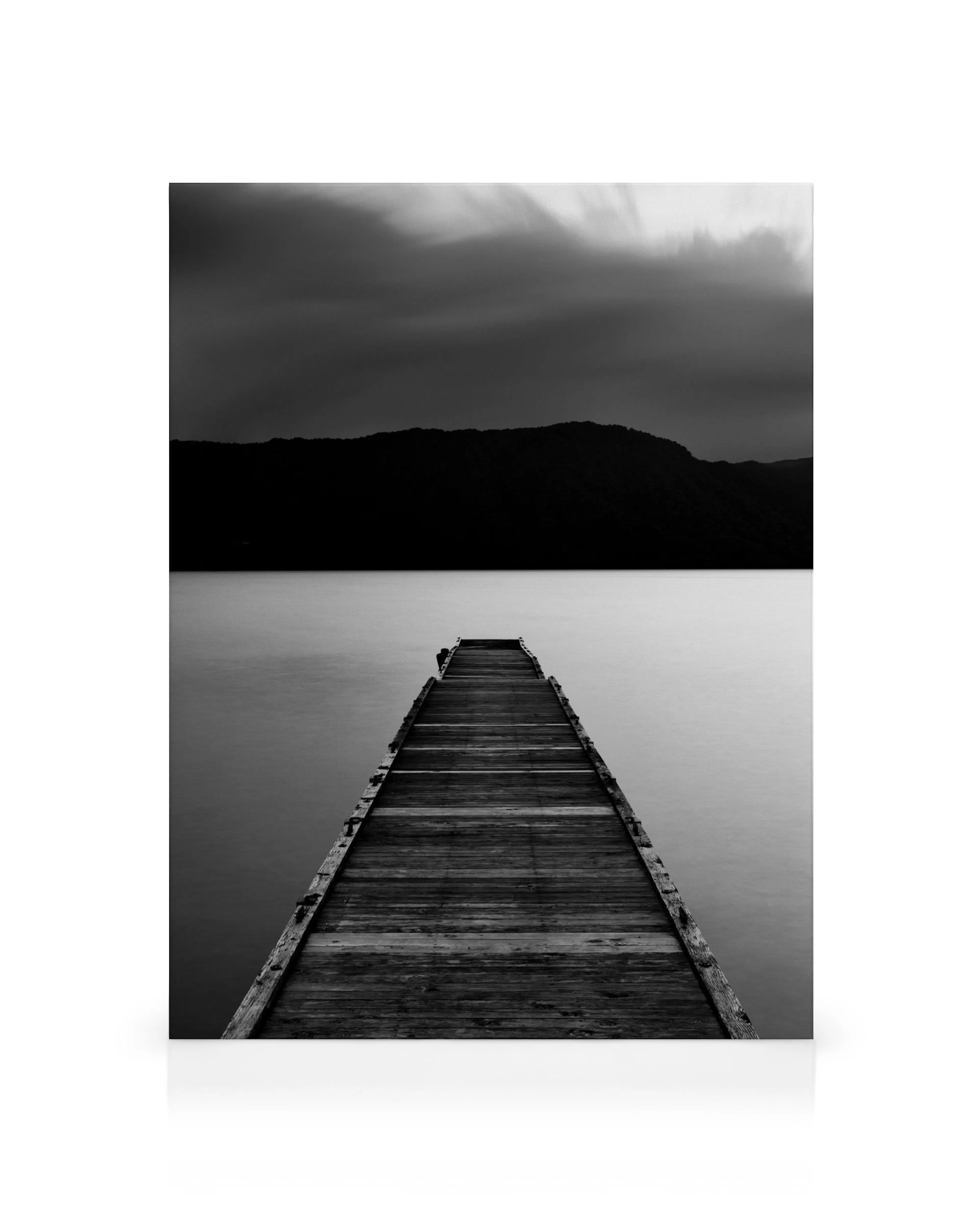Black and white canvas print of a wooden pier over calm water leading to distant hills under a cloudy sky.