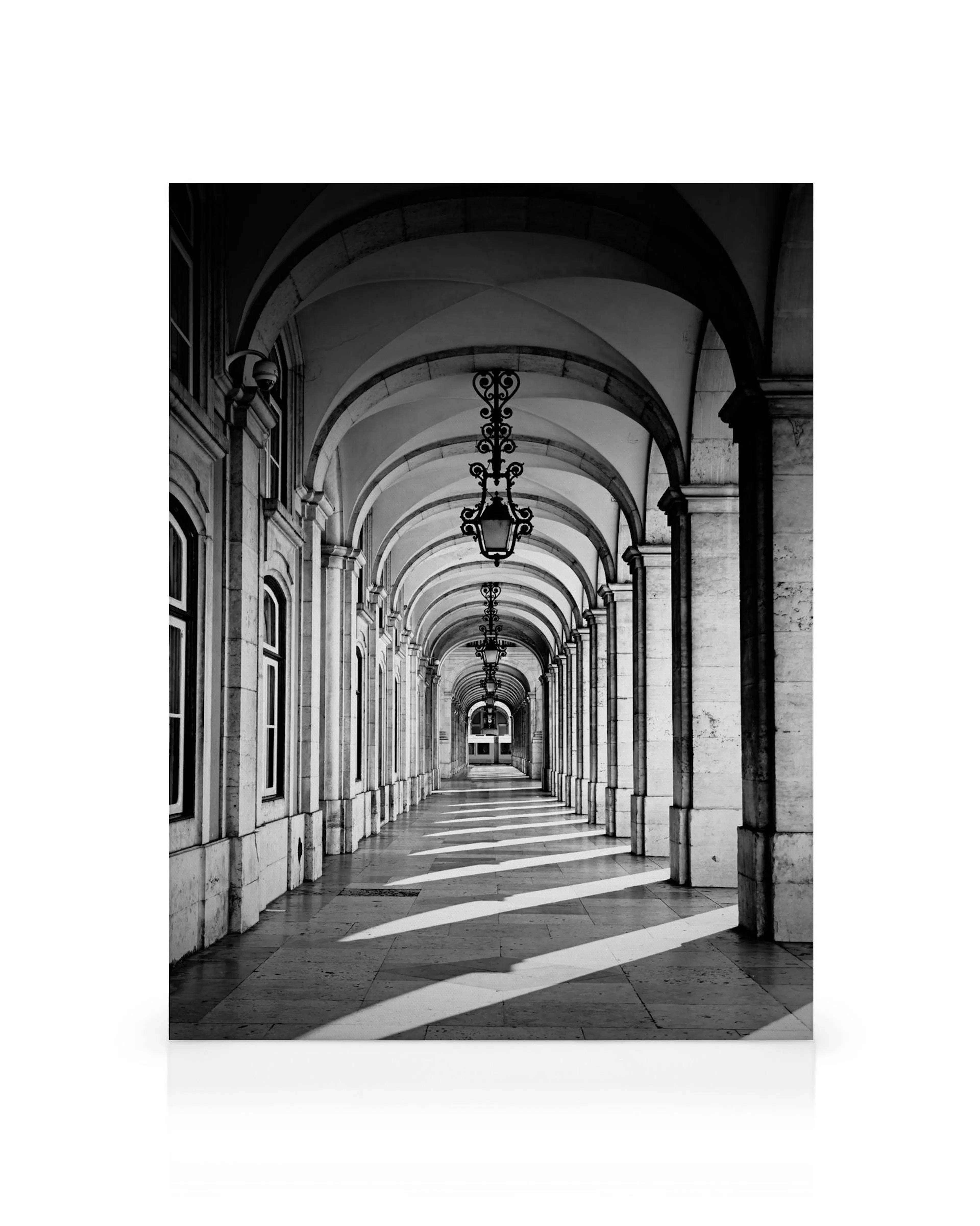 Black and white photograph of a long arched corridor with ornate hanging lamps, sunlight casting dramatic shadows on the floor.