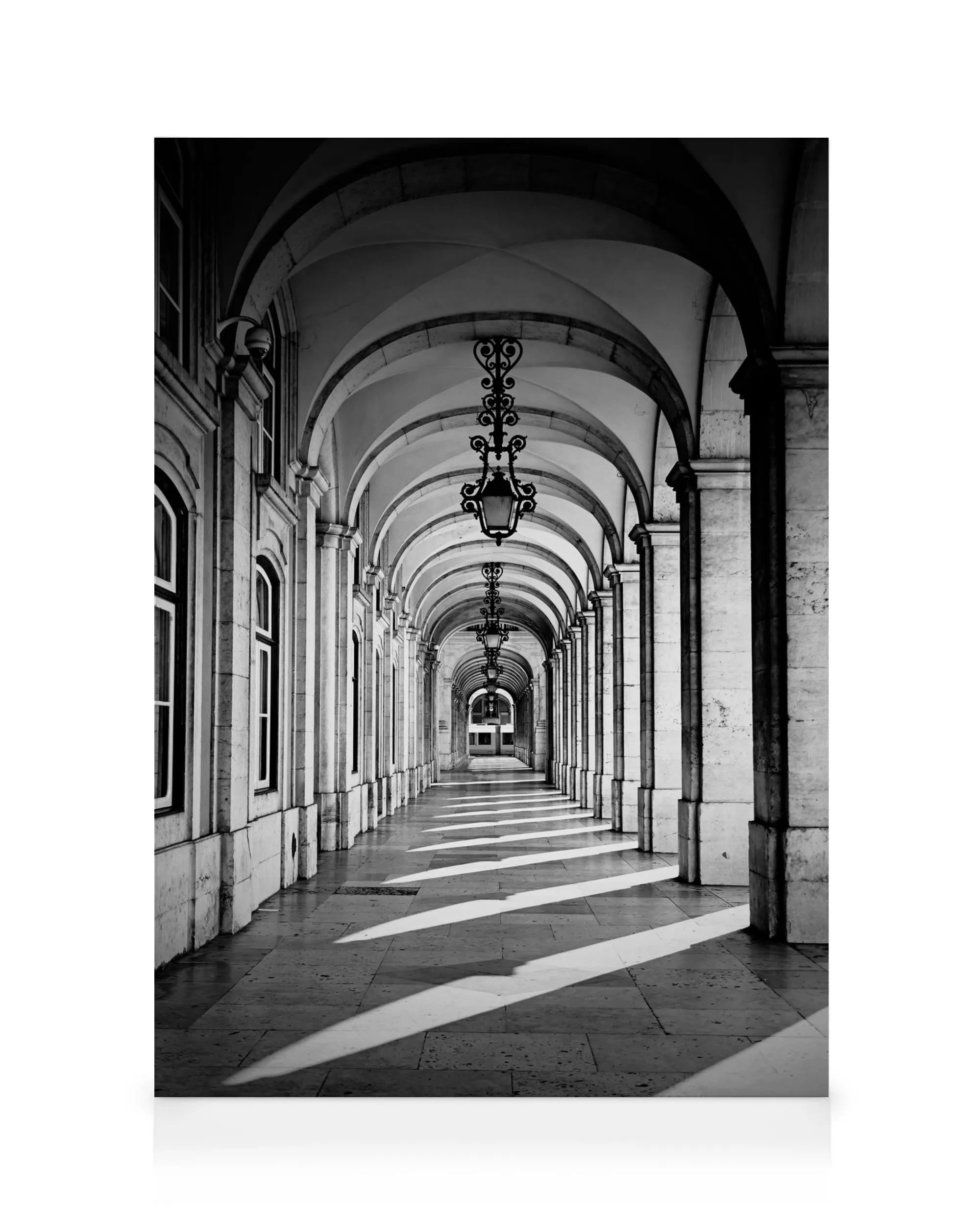 Black and white photograph of a long arched corridor with ornate hanging lamps, sunlight casting dramatic shadows on the floor.