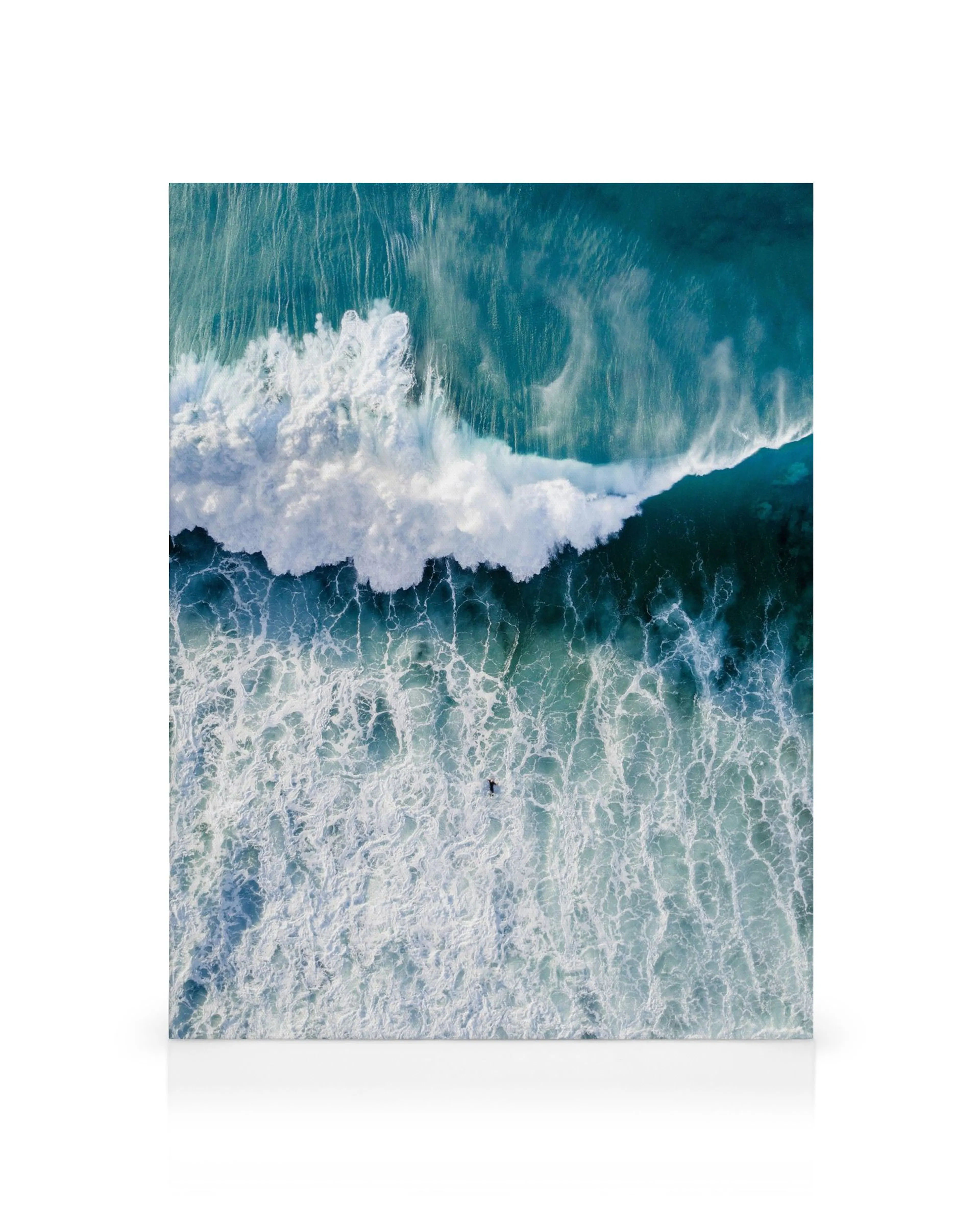Aerial view of a teal ocean wave breaking with white foam, a lone surfer in the water below.