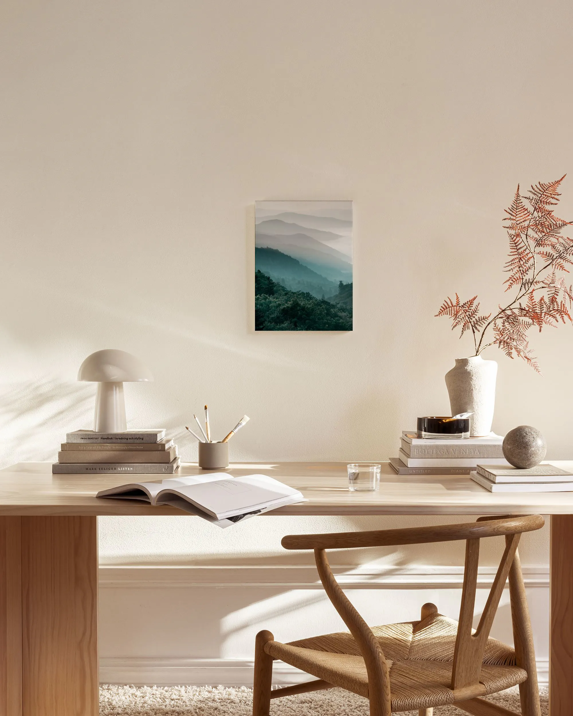 A canvas print of misty teal mountains with dark green trees, hanging in a bright, minimalist living room above a wooden console
