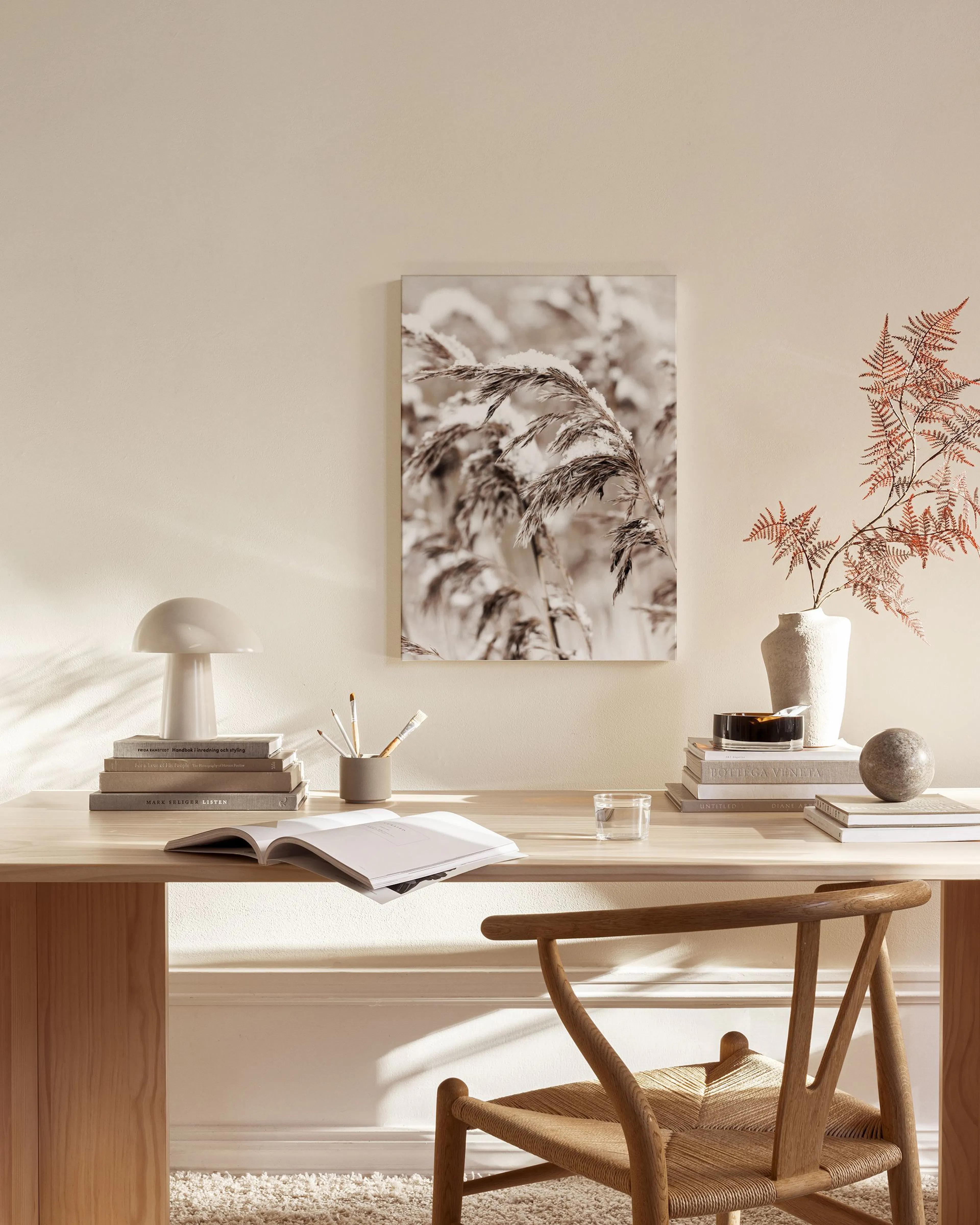Een canvas met sepia-getinte rietpluimen, hangend boven een houten dressoir met lamp en boeken in een lichte woonkamer.