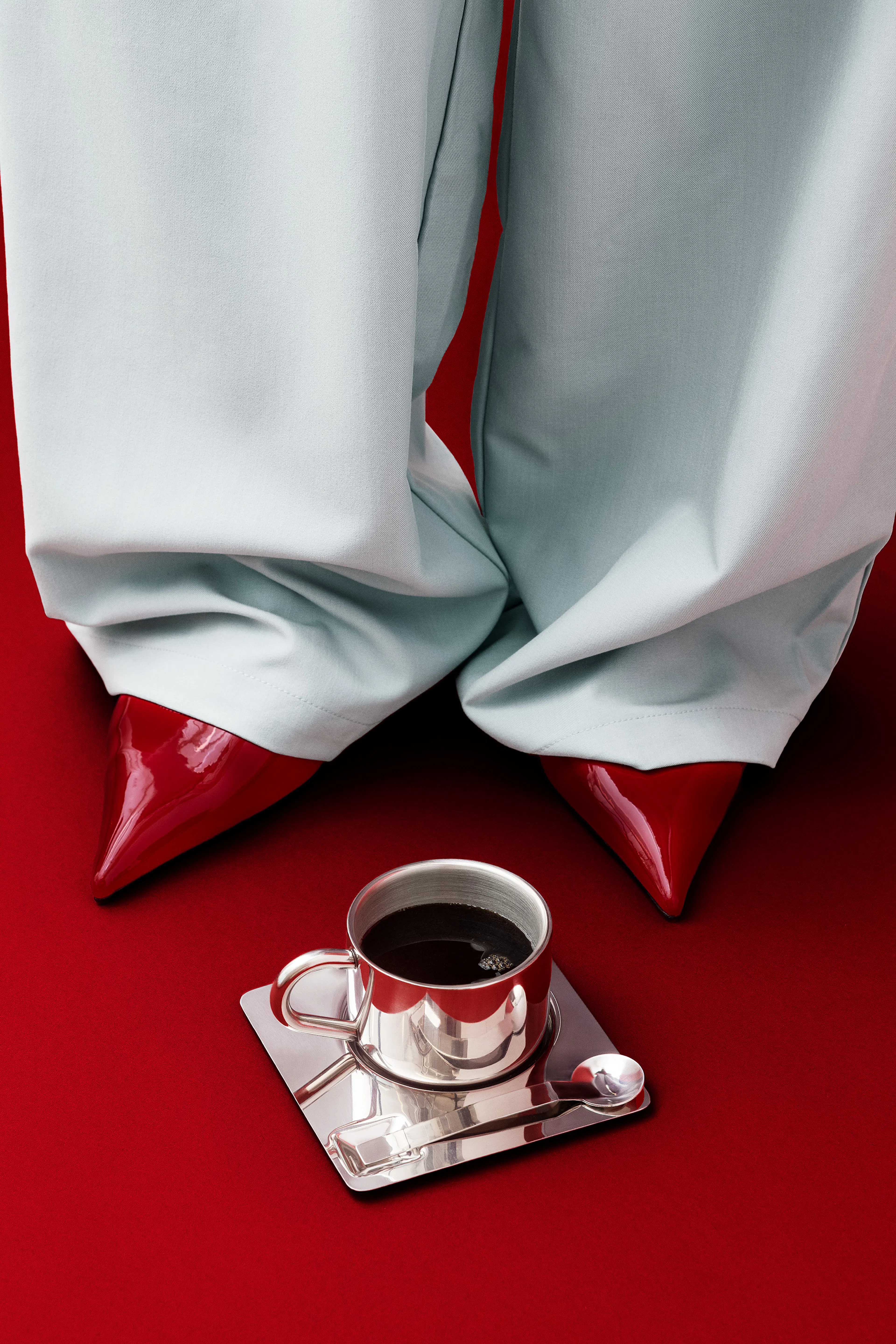Poster con gambe in pantaloni chiari e scarpe rosse lucide su sfondo rosso, con una tazza dargento di caffè.