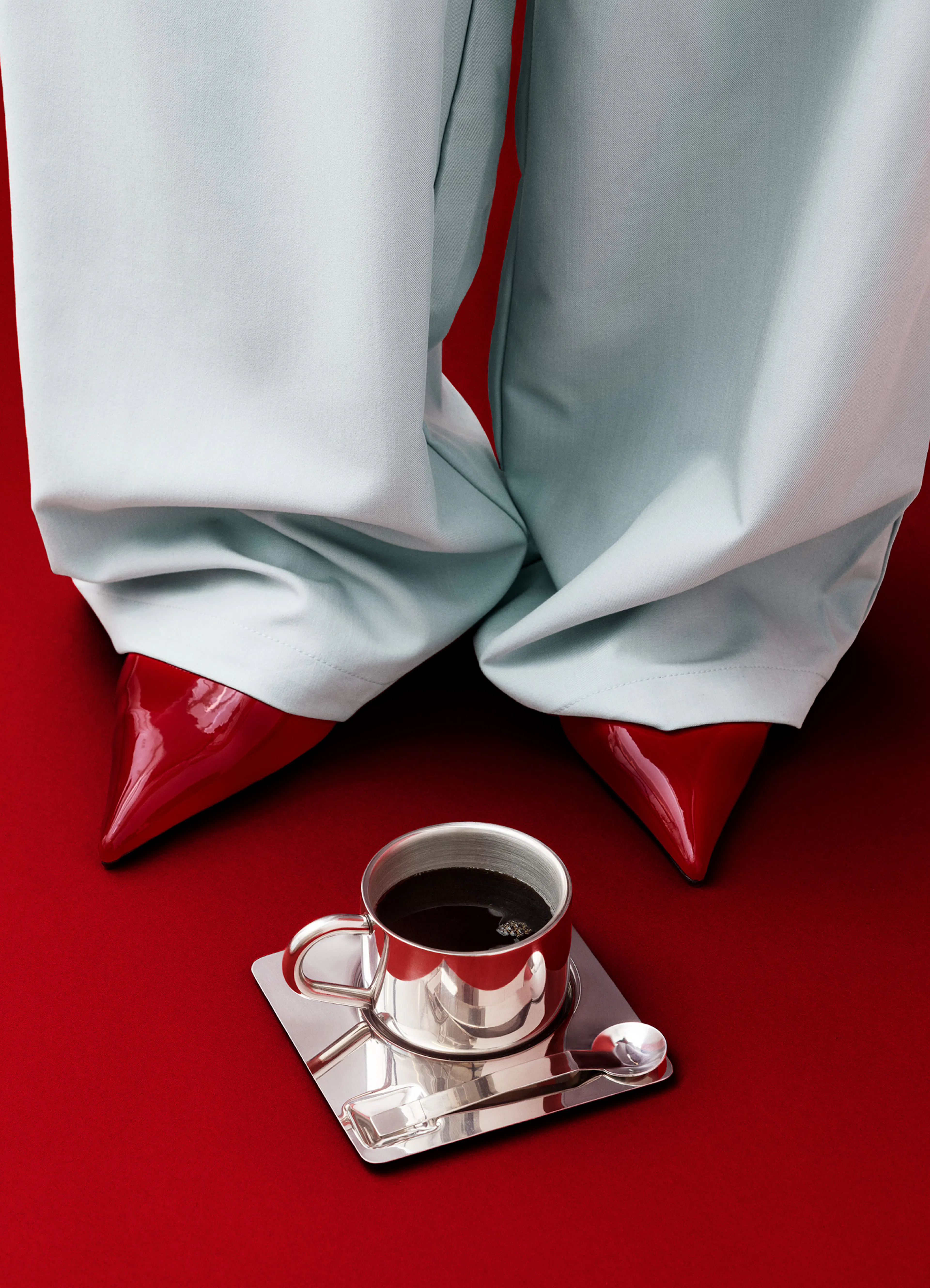 Poster con gambe in pantaloni chiari e scarpe rosse lucide su sfondo rosso, con una tazza dargento di caffè.