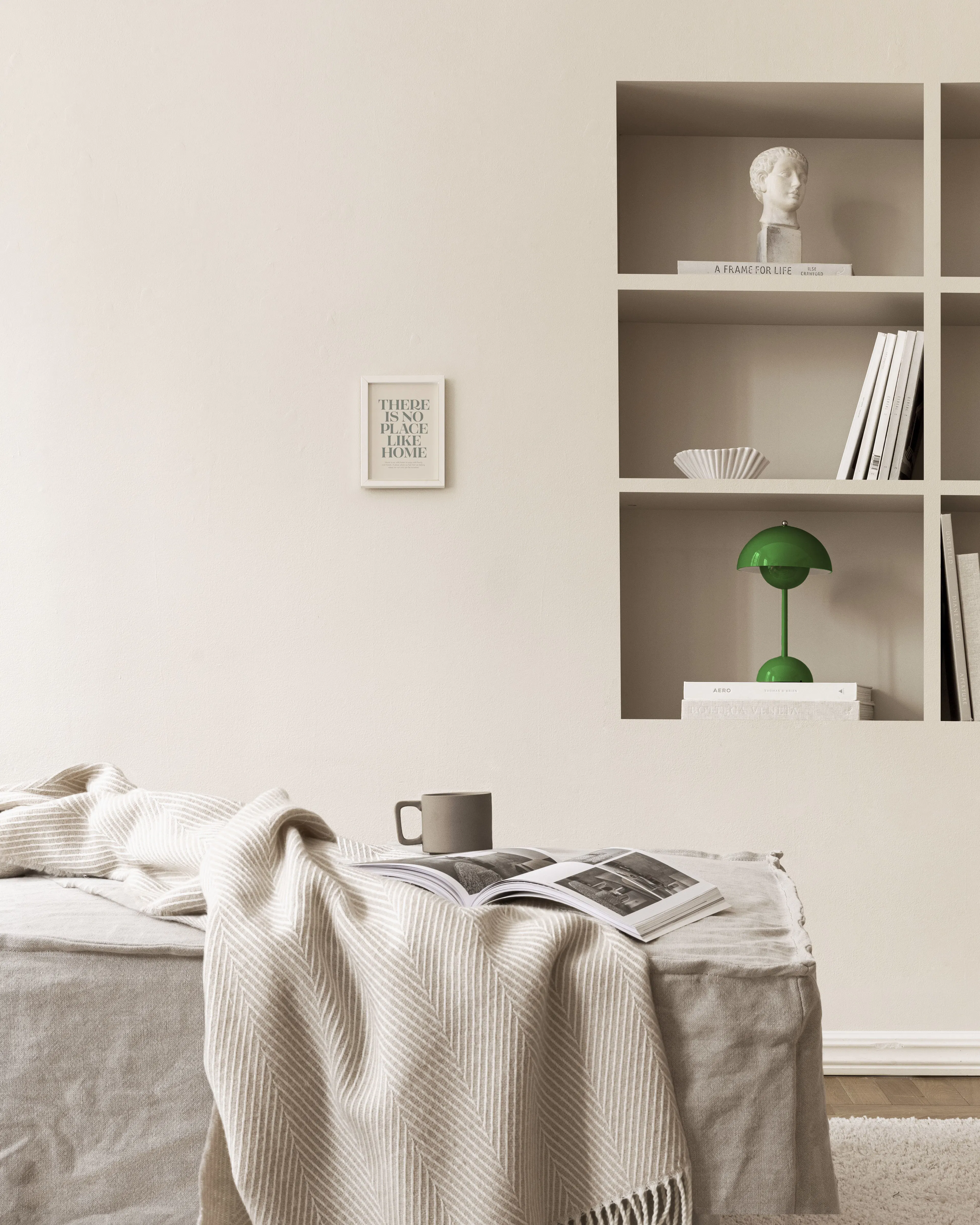 Plakát s nápisem There Is No Place Like Home v béžových tónech, visící nad pohovkou s přehozem a knihou v béžovém obývacím pokoj