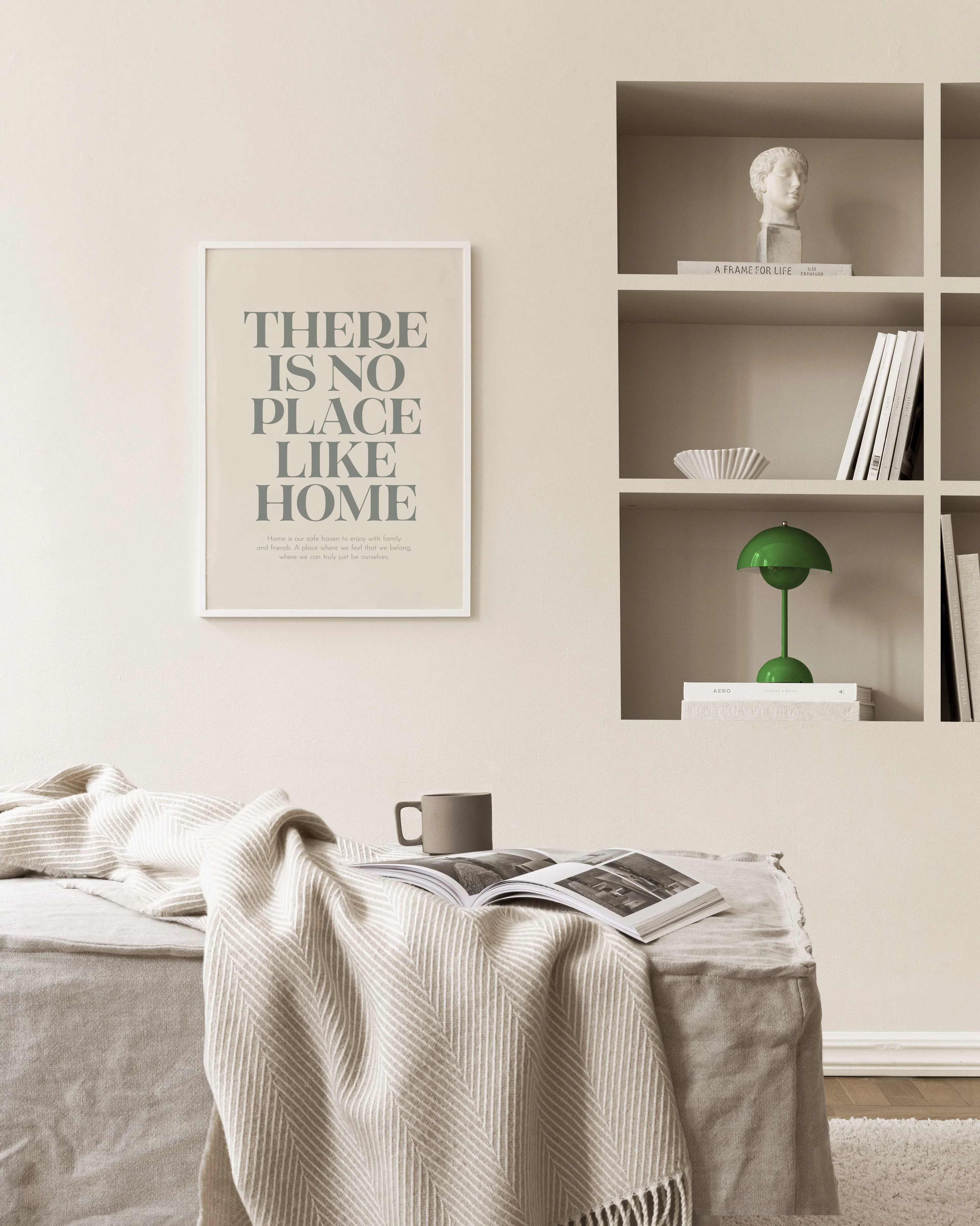 A print with the quote There is no place like home in grey text, displayed above a beige sofa with a mug and book.