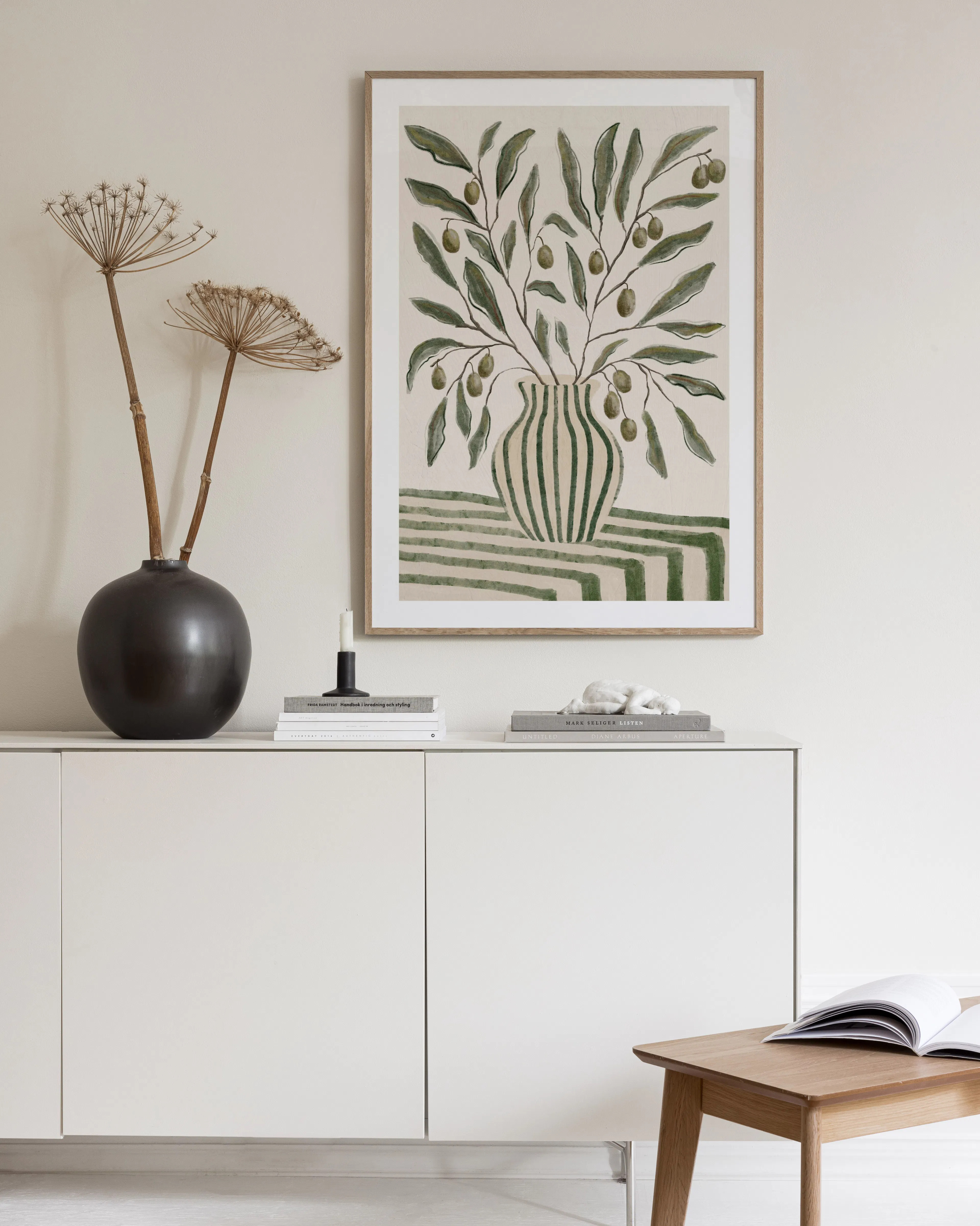 A print of an olive tree branch in a striped vase on a striped surface, hanging above a wooden desk.