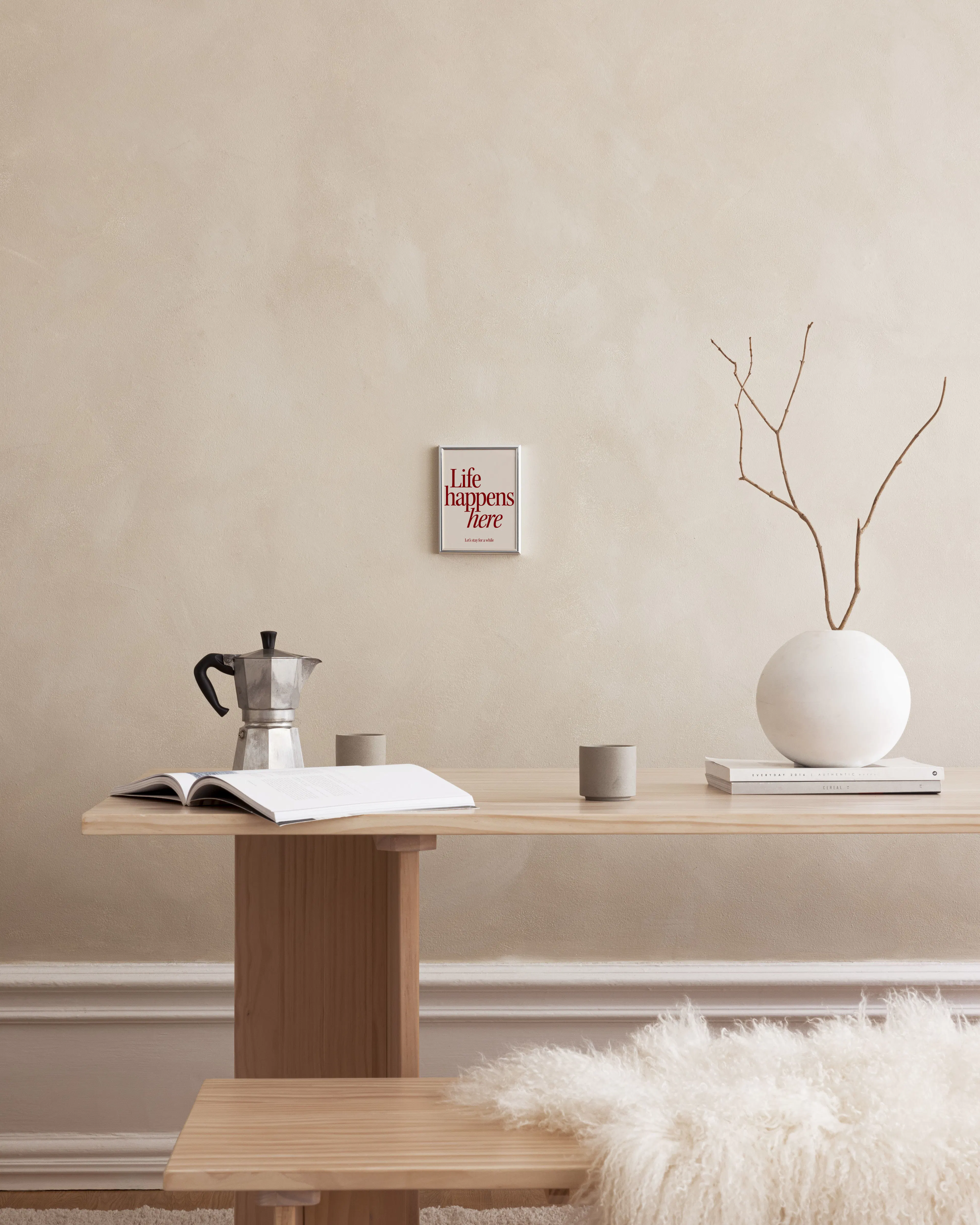 Poster mit dem Text Life happens here in Rot, hängt über einem Holztisch mit Kaffeekanne und Vase.
