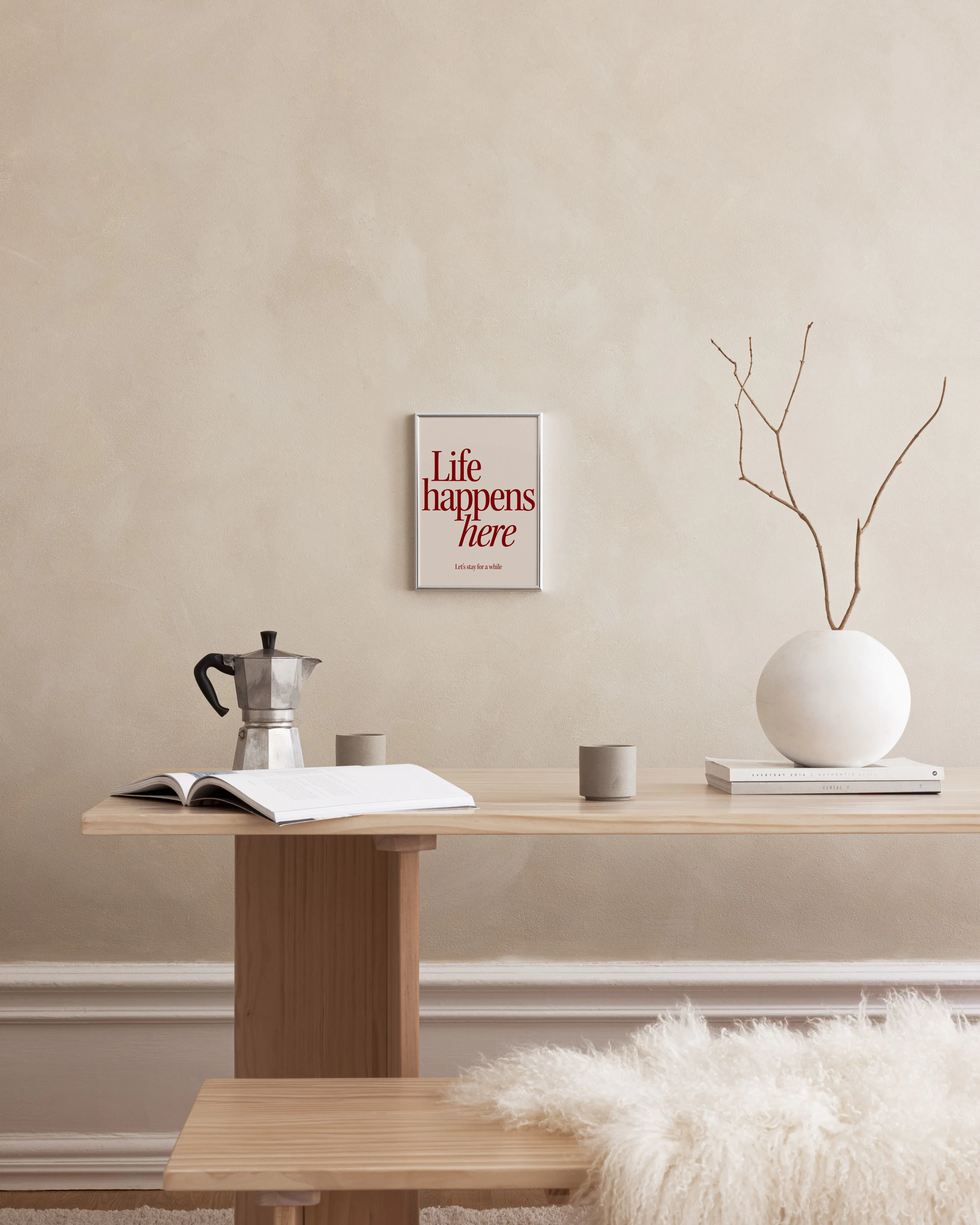 Plakat med rød tekst Life happens here på beige baggrund, ophængt over et træbord med en bog, kaffekande og vase.