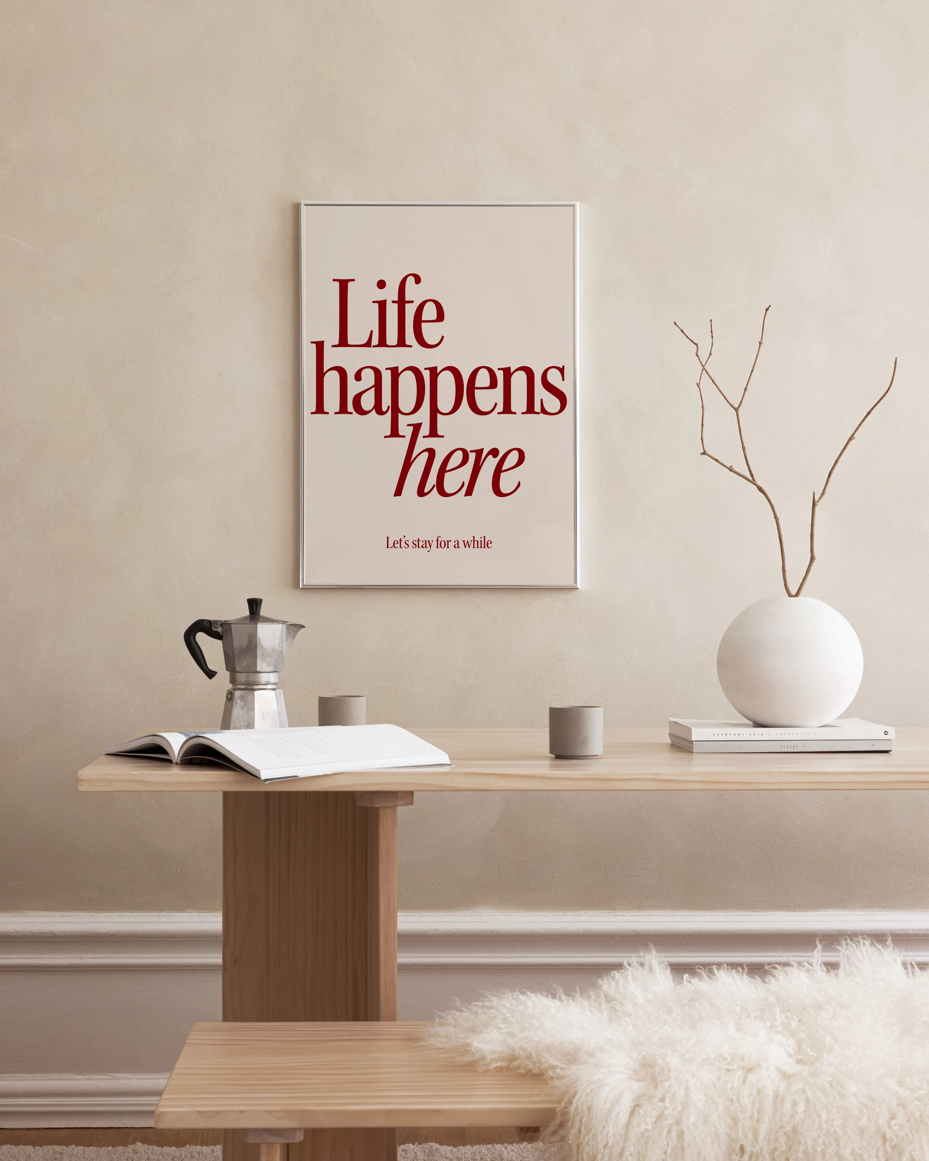 Póster Life happens here con letras rojas sobre fondo beige, colgado sobre una mesa de madera con cafetera y jarrón.