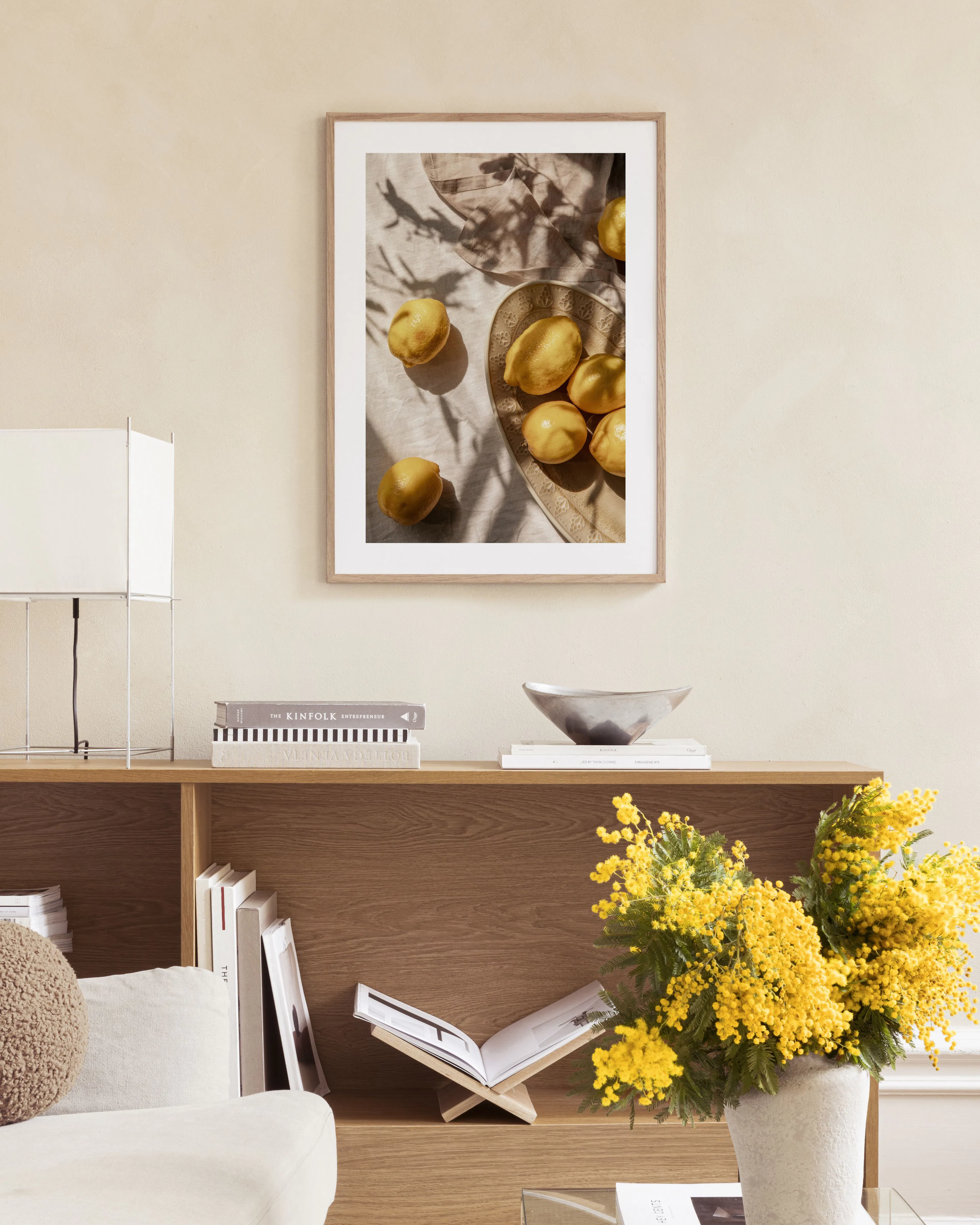 A print of lemons on a beige linen tablecloth with shadows, hanging above a natural wood dresser next to a lamp.