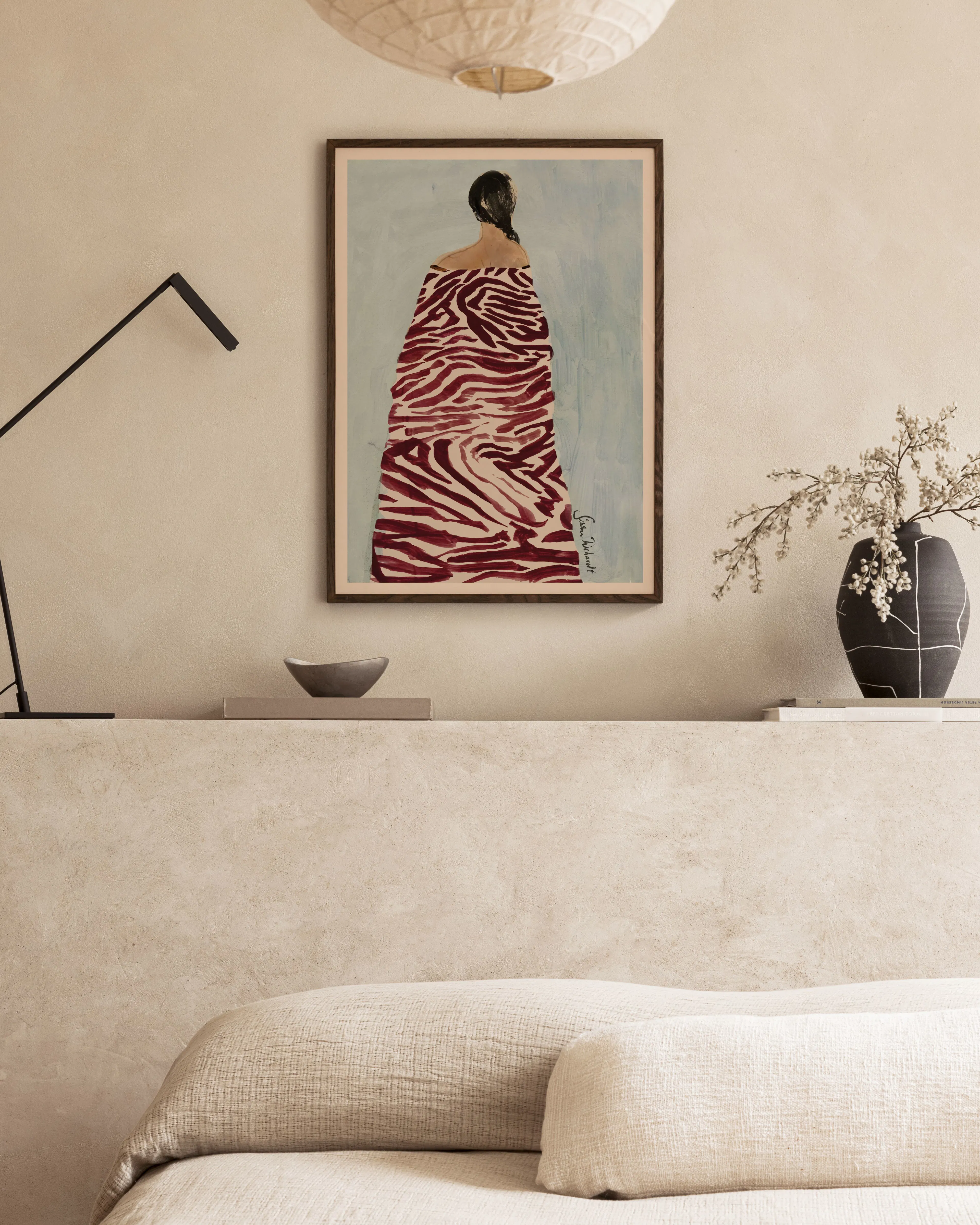 Póster de una figura femenina vestida de rojo y blanco, exhibido en una repisa de sala con una lámpara negra y un jarrón.